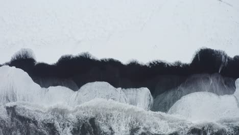 Eine-Wunderschöne-Drohnenaufnahme-Des-Schwarzen-Strandes-In-Island-Zeigt-Die-Hohen-Wellen-Des-Meeres-Und-Das-Bewölkte-Wetter,-Während-Der-Schnee-Einen-Teil-Des-Schwarzen-Sandes-Bedeckt
