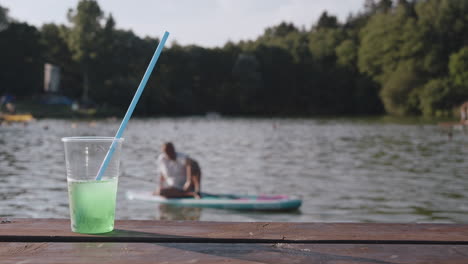 Drink-in-plastic-cup-with-straw-and-person-on-paddleboard-in-background,-summer-time-or-holiday-on-lake