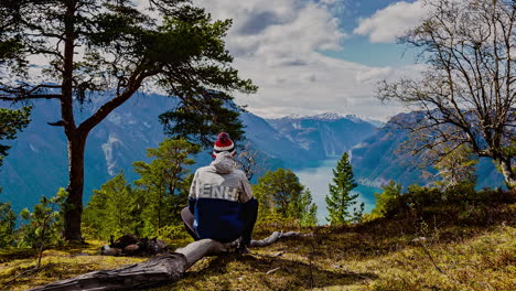 Shot-of-a-young-man-sitting-on-a-dead-tree-and-enjoying-the-scenic-view-fjord-along-beautiful-mountain-range-in-Norway-on-a-cloudy-day