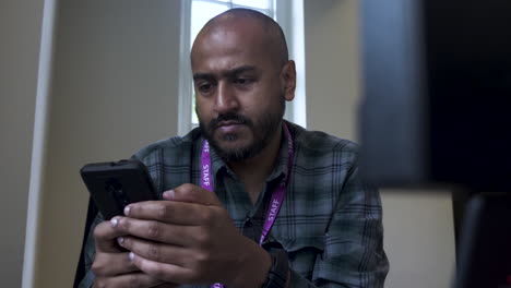 A-close-up-shot-from-behind-an-office-computer-as-an-Asian-Indian-man-sits-at-his-desk-responding-to-a-text-message-on-his-mobile-phone