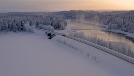 Aerial-view-orbits-freezing-Scandinavia-woodland-lake-crossing-following-vehicle-on-journey-through-Lapland-wilderness