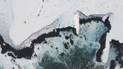 A-beautiful-drone-shot-of-the-Beach-in-Iceland-shows-the-high-waves-of-the-sea-and-the-cloudy-weather-while-the-snow-covers-some-of-the-black-sand