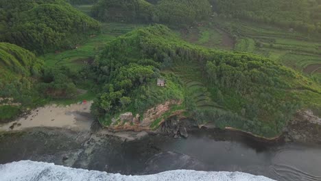 Orbit-drone-shot-of-waves-reaching-green-coastline-of-Indonesia-with-private-house-on-cliff