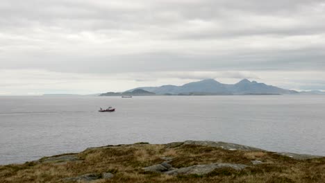 Slow-panning-shot-looking-out-to-sea-as-a-red-fishing-boat-moves-in-front-of-mountains-on-Scottish-islands-in-the-background-and-heather-moorland-in-the-foreground