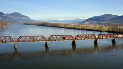 Mission-Railway-Bridge-With-Freight-Train-Crossing-In-Mission,-British-Columbia,-Canada---aerial-drone-shot