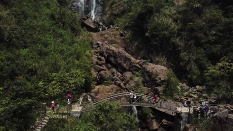 A-fast-drone-shot-reverses-to-reveal-a-transition-from-a-waterfall-to-tourists-waving-from-a-walkway-bridge
