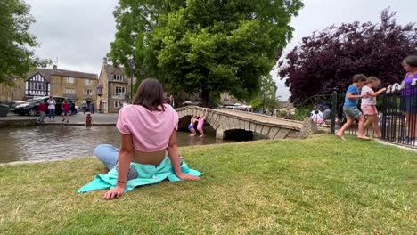Girl-walking-the-Cotswolds-Way-relaxs-in-the-grass-by-the-riverside-in-Bourton-On-The-Water,-Cotswolds---England,-UK