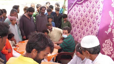 Lugareños-De-Pie-Alrededor-De-Un-Trabajador-Humanitario-Con-Mascarilla-Escribiendo-Detalles-De-Los-Refugiados-De-Las-Inundaciones-En-El-Campamento-De-Socorro-En-Sindh