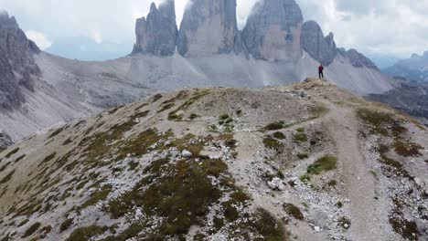 A-cool-4K-drone-shot-of-a-hiker-standing-on-a-mountain-top-surrounded-by-the-impressive-rock-formations-and-mountain-landscape-of-the-Dolomites-in-Italy---Looking-at-the-stunning-Tre-Cime-di-Lavaredo