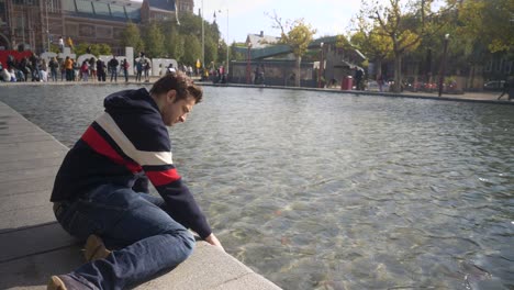 Handsome-young-man-sitting-on-the-bank-of-the-canal-in-front-of-Rijksmuseum-with-I-Amsterdam-sign-putting-hand-in-the-water