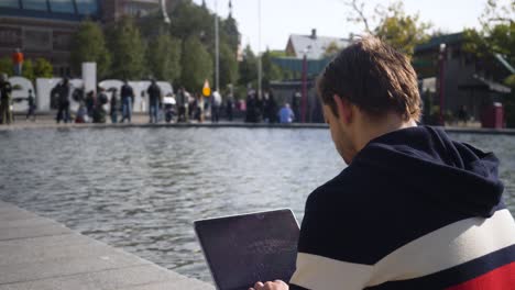 Nice-looking-young-man-sitting-at-the-museumplein-in-Amsterdam-typing-on-the-laptop-in-a-sunny-day