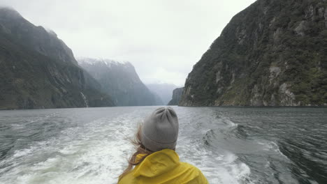 Mädchen-In-Einer-Leuchtend-Gelben-Regenjacke,-Die-Auf-Der-Rückseite-Eines-Bootes-Steht,-Während-Es-An-Einem-Regnerischen-Und-Kalten-Tag-Durch-Die-Fjorde-Neuseelands-Fährt