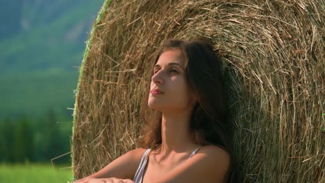 A-beautiful-brunette-woman-sitting-calmly-in-thought-against-a-large-hay-bale-with-a-straw-of-grass-in-her-mouth-as-she-ponders