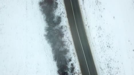 Birds-eye-view-of-empty-road-in-winter,-landscape-covered-in-snow,-black-car-driving-on-the-road,-anticipation-of-something-to-happen
