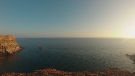 Sunset-in-TiritaÃ±a-Beach,-Gran-Canaria,-Spain,-with-a-boat-sailing-and-blue-sky-framed-by-high-cliffs-near-Taurito-Mogas,-between-Punta-de-la-Cruz-de-Piedra-and-Punta-de-los-Medios-Almudes
