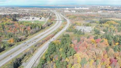 Aerial-footage-facing-towards-Ottawa,-Ontario-with-a-4-lane-highway-to-the-left-and-buildings-in-the-distance-and-autumn-forests-on-either-side