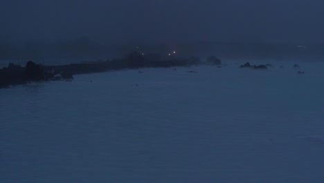 Islandia,-Laguna-Azul,-Estación-De-Energía-Geotérmica-Svartsengi-En-La-Noche,-Figura-Distante-Caminando-Por-Un-Sendero-Sobre-El-Agua,-Plano-General