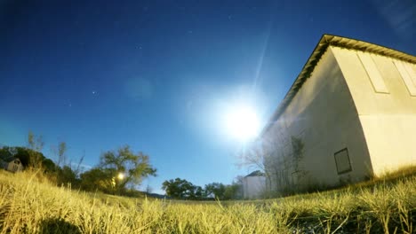 NIGHT-LAPSE---Blue-sky-with-the-moon-and-stars-flying-by-a-field-with-an-old-abandoned-building