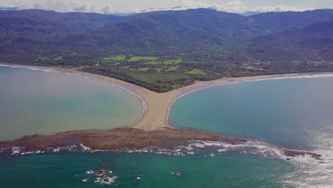 Aerial-shot-pulling-away-from-the-whale-tail-shaped-rocky-point-of-Punta-Uvita-surrounded-by-bright-blue-ocean-waves-and-lush-rainforest-in-the-distance-in-south-Puntarenas,-Costa-Rica
