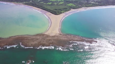 Aerial-shot-pulling-away-from-the-whale-tail-shaped-rocky-point-of-Punta-Uvita-surrounded-by-bright-blue-ocean-waves-in-south-Puntarenas,-Costa-Rica