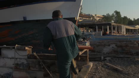 Painter-steps-up-to-paint-Greek-fishing-boat-real-time-handheld-shoreline-harbour-sunshine