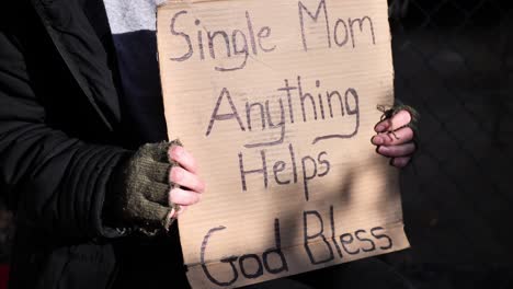 Homeless-women-sitting-down-holding-cardboard-sign-reading,-"Single-mom,-anything-helps,-God-bless"