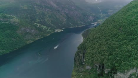 Slomo-Luftaufnahme-Des-Geirangerfjords,-Norwegen,-Mit-Einem-Fahrenden-Boot-Und-Einer-Stadt-Im-Hintergrund