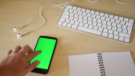 beautiful-female-hand-scrolling-and-tapping-on-phone-with-green-screen-on-a-minimal-workplace-with-keyboard,-earphones-and-notebook