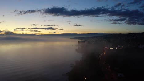 Aerial-shot-descending-towards-lake-shore-through-mist,-at-sunset-with-patches-of-fog-reflecting-in-the-water-Lutry,-Vaud---Switzerland