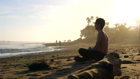 A-muscular-man-watching-the-sunset-in-a-meditation-pose-as-surfers-walk-along-the-ocean-water-in-silhouette-in-golden-sunlight