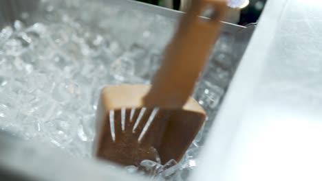 Close-up-of-a-metal-container-with-ice-cubes-and-a-gold-ice-scoop-in-it,-inside-a-bigger-bin-with-glass-alchol-bottles-inside