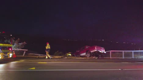 Pan-across-roadside-accident-scene,-firemen-walk-around-car-crashed-through-fence