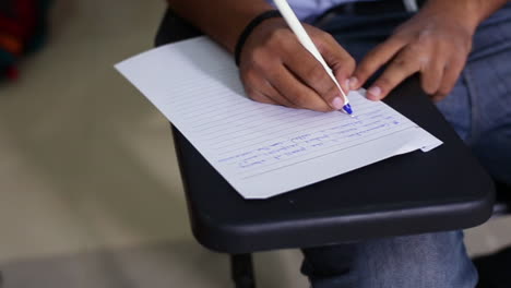 A-top-close-up-view-of-a-boy-student-writing-on-the-paper-at-his-class-room,-wearing-a-jeans-pant,-A-chair-with-its-writing-desk