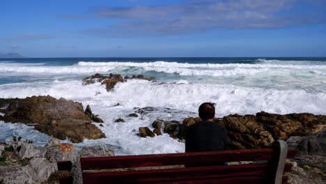 Pensive-man-on-bench-overlooking-ocean-and-waves-breaking-onto-rocky-coastline