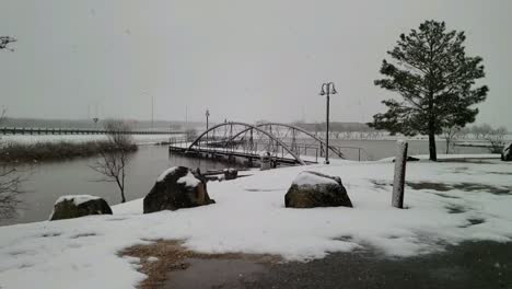 Watching-the-snow-fall-in-slow-motion-on-small-Texas-city-lake-with-beautiful-walking-bridge,-trees,-lake,-rocks-and-gray-skies
