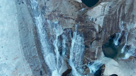 Aerial-top-view-ascending,-revealing-cascading-waterfalls-into-deep-blue-pools-in-granite