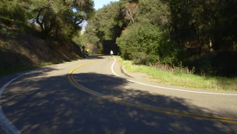 2-motorcycles-drive-down-a-windy-road-between-green-trees-in-California-as-a-single-man-walks-in-the-background-alone