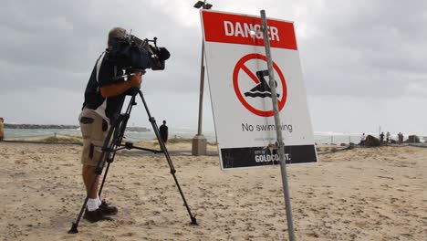 Ein-Nachrichtenkameramann-Neben-Einem-Warnschild,-Das-Videoaufnahmen-Von-Schäden-An-Einem-Strand-Durch-Einen-Sich-Entwickelnden-Zyklon-Macht