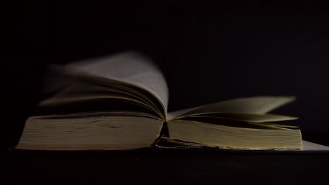 Slow-motion,-close-up-of-an-old,-vintage,-open-book-with-pages-turning-on-the-wind,-with-glowing-sparkles-flying-around,-on-a-dark-room-with-few-light,-with-black-surroundings