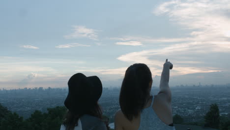 Two-Teenage-Girls-Taking-A-Photo-And-Pointing-On-The-Sky-Of-A-City-Sunset