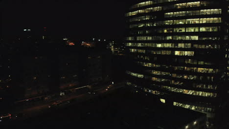 Aerial-night-shot-of-a-tall-office,-financial-building-as-people-working-late-with-lights-on-in-the-offices,-light-traffic-on-the-roads