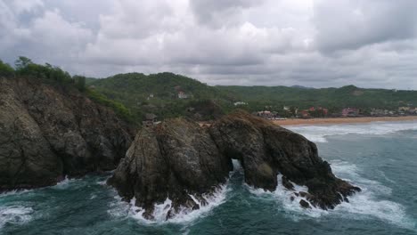 Aerial-jib-down-shot-of-a-big-rock-formation-with-a-door-in-the-middle,-Zipolite-beach,-Oaxaca