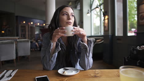 Attractive-latina-brunette-sitting-at-the-table-having-a-coffee,-waiting,-thinking-and-looking-outside