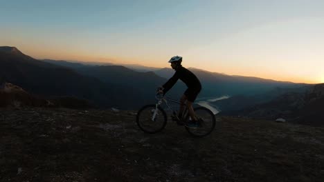 Man-riding-a-bike-at-the-top-of-a-mountain-with-beautiful-canyon-lake-at-sunset-in-the-background