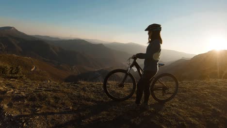 Chica-Empujando-Bicicleta-En-La-Cima-De-Una-Montaña-Al-Atardecer