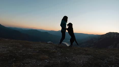 Girl-playing-and-hugging-with-black-labrador-dog-on-a-mountain-at-sunset