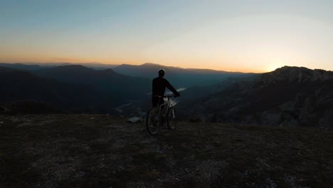Young-man-standing-with-his-bike-at-a-top-of-a-mountain-at-sunset