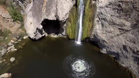 Aerial-Drone-shot-of-male-jumping-off-cliffs,-at-waterfall-splashes-into-water-while-female-watches-from-above