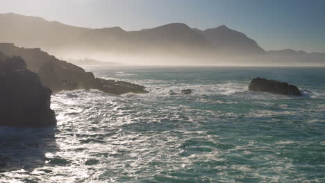 Early-morning-mist-over-ocean,-as-sun-rises-and-waves-run-onto-rocky-coastline,-Hermanus,-South-Africa