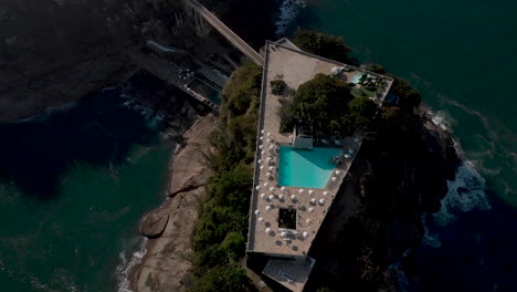 Top-down-aerial-view-of-a-recreational-construction-on-a-small-island-on-the-coast-of-Rio-de-Janeiro-seen-from-above-at-sunset-with-umbrellas-casting-a-shade-around-the-pool